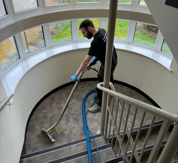 A professional cleaner using a carpet cleaning machine on a curved stairwell landing, with large windows providing natural light and a view of the greenery outside.