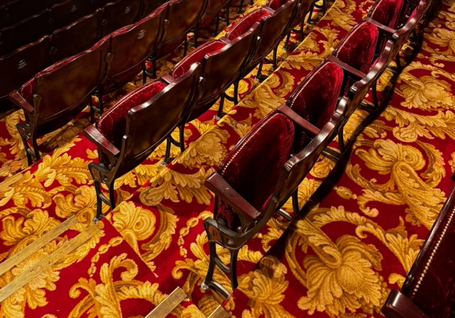 Rows of empty, dark red theatre seats with wooden backs, set on a bright red carpet with an intricate gold floral pattern.