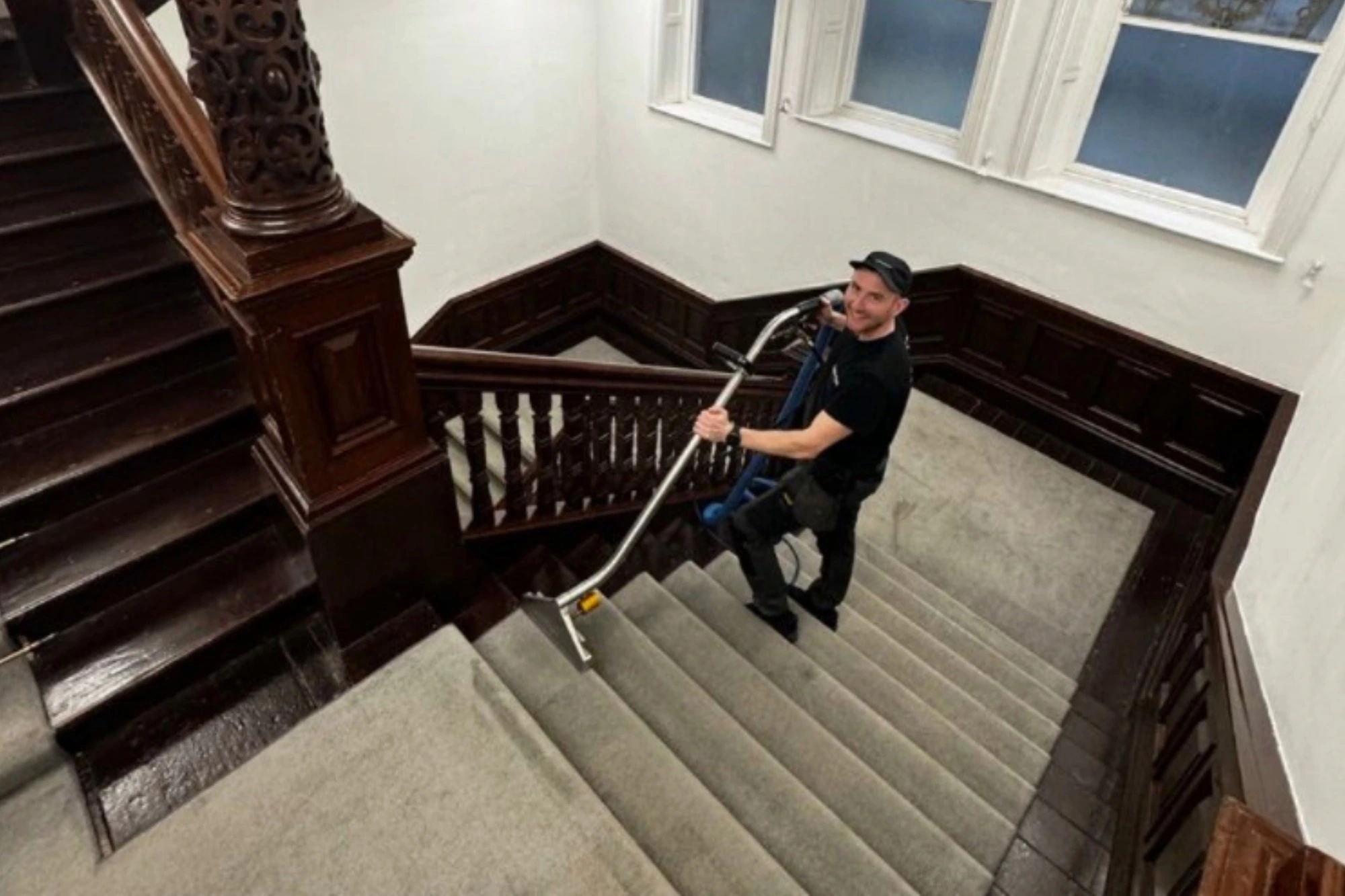Harry Dyson using professional carpet cleaning equipment on a grand wooden staircase with stained-glass windows in the background.