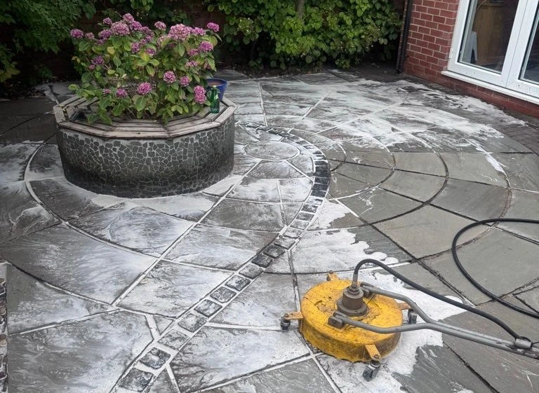 A patio being cleaned using a yellow rotary surface cleaner on wet, grey flagstones.