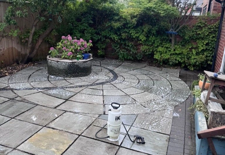 A wet, circular stone patio being treated with cleaning foam, with a white pump sprayer in the foreground and a stone flower planter in the centre.