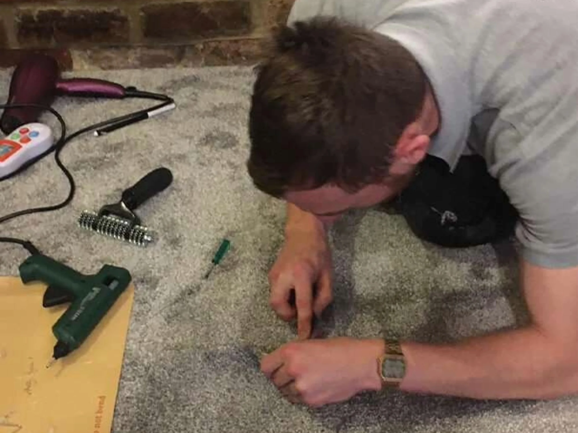 A person works on repairing a grey carpet, surrounded by tools like a hot glue gun, a roller, and a hairdryer near a brick wall.