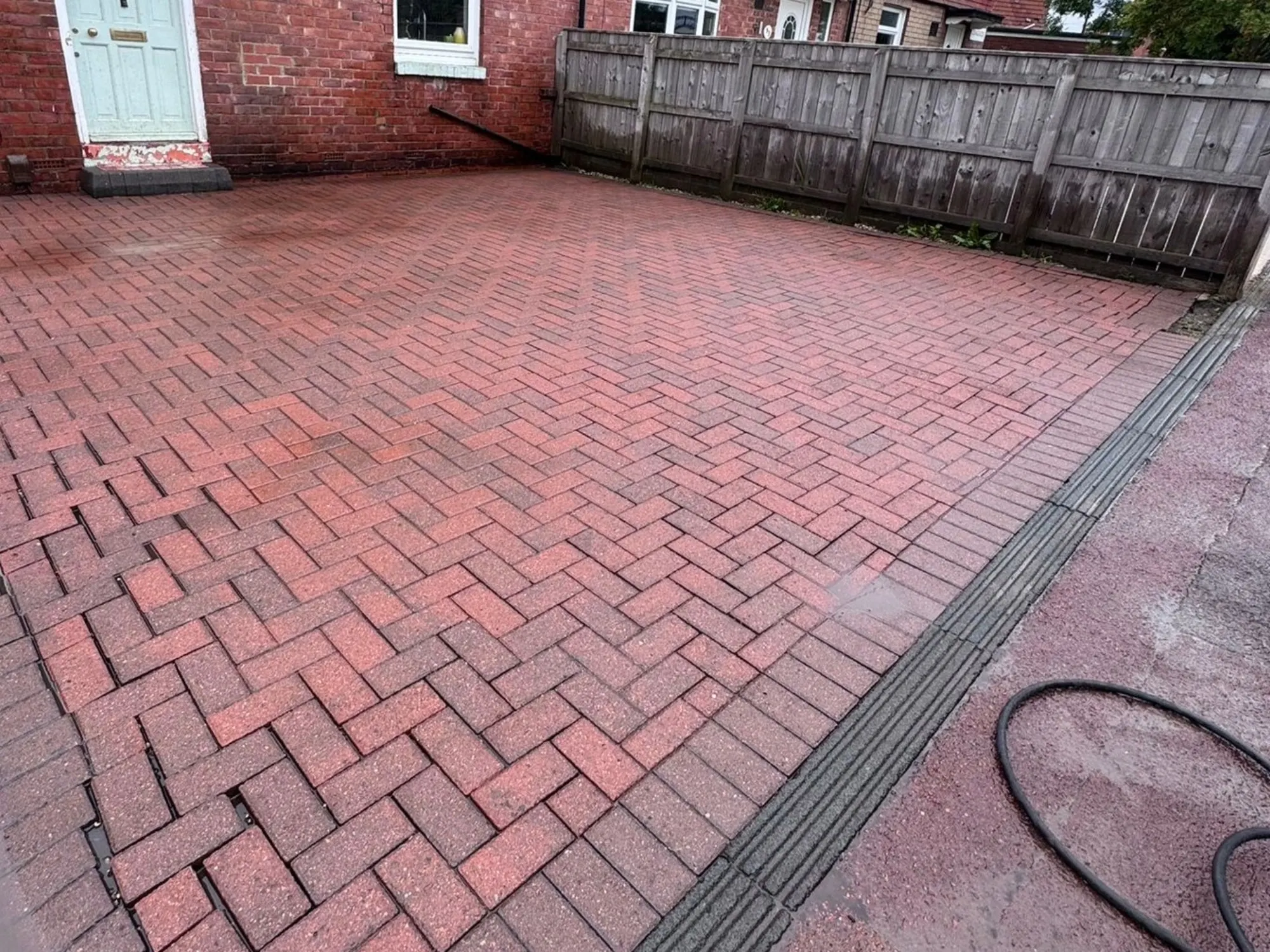 A red block-paved driveway in a herringbone pattern alongside a red brick house and a grey wooden fence.