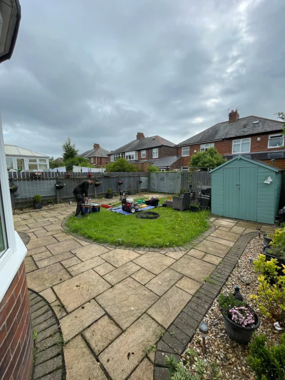 A garden with a small circle of grass surrounded by paving slabs that have crumbling pointing filled with dirt and debris.